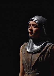 A person stands under a spotlight against a dark background, wearing a rough, sleeveless garment with a shiny, metallic hood. The expression is thoughtful and introspective.