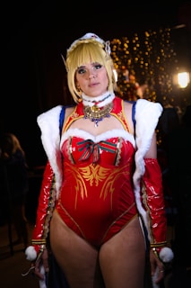 A person is dressed in an elaborate red and gold costume with white fur accents and intricate jewel details. The background features a softly lit setting with fairy lights.
