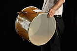 A person holding a large, wooden drum with their left hand, while their right hand holds a drumstick. The drum is positioned horizontally, and the image captures a partial view of the person dressed in a white shirt and black pants.