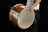 A person holding a large, wooden drum with their left hand, while their right hand holds a drumstick. The drum is positioned horizontally, and the image captures a partial view of the person dressed in a white shirt and black pants.