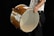 A person holding a large, wooden drum with their left hand, while their right hand holds a drumstick. The drum is positioned horizontally, and the image captures a partial view of the person dressed in a white shirt and black pants.