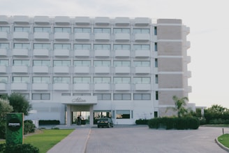 A modern, multi-story hotel building with a symmetrical and clean facade featuring rectangular windows and small balconies. The entrance is clearly marked, and there are signs of a well-maintained garden with a variety of plants and trees around the perimeter. A parked car is visible near the entrance, and a sign labeled 'Ipanema' is present on the left side of the image.