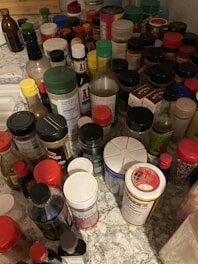 A cluttered kitchen countertop filled with various spices and condiments. There are numerous bottles, jars, and containers of different sizes, shapes, and colors, including some recognizable brands of sauces and seasonings. The items are spread out across a marble-like counter space.