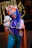 A person dressed in an elaborate and colorful costume inspired by a clown, featuring a bright orange hat adorned with a skull emblem, a blue wig, and a red nose. The individual is smiling broadly and holding a plush toy character. The costume includes a striped shirt, blue pants, and an orange cape with feather accents. The background is decorated with twinkling lights, adding to a festive atmosphere.