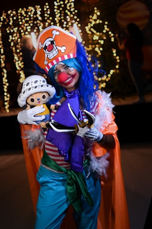 A person dressed in an elaborate and colorful costume inspired by a clown, featuring a bright orange hat adorned with a skull emblem, a blue wig, and a red nose. The individual is smiling broadly and holding a plush toy character. The costume includes a striped shirt, blue pants, and an orange cape with feather accents. The background is decorated with twinkling lights, adding to a festive atmosphere.