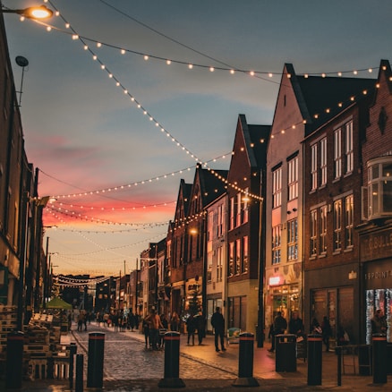 A quaint street scene at dusk featuring charming old-style buildings with steep gabled roofs. String lights are beautifully draped across the street, casting a warm glow over the cobblestone path. The sky is a mix of soft pink and blue hues as the sun sets. People are leisurely walking and shopping, adding a lively atmosphere to the scene.