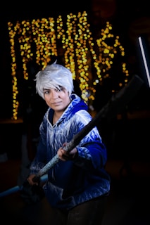 A person with white hair is dressed in a blue hoodie with icy patterns, holding a long staff, standing confidently. The background is adorned with glistening fairy lights creating a vibrant, magical atmosphere.