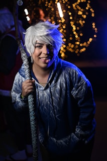 A person with white hair wearing a blue, winter-themed hoodie and holding a staff. The individual is sticking their tongue out and appears to be at a festive event with string lights in the background.