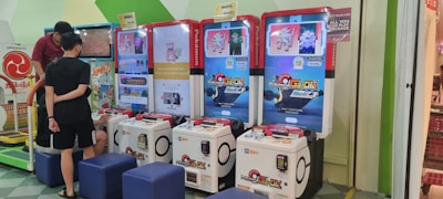 Several arcade gaming machines with multiple screens displaying Pokémon-themed content are positioned in a colorful arcade setting. There are people interacting with the machines; some are seated, and others are standing. The area features different gaming options, including a jumping game with bright, inviting graphics.