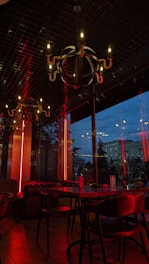 A dimly lit restaurant interior with modern chandeliers hanging from a dark ceiling. The walls have large glass windows showing an evening view outside. Red neon lights add a warm yet intense ambiance to the setting, reflecting off the wooden furniture and creating a cozy atmosphere.