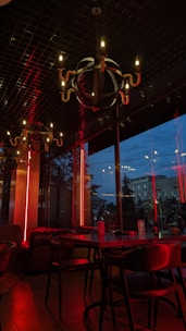 A dimly lit restaurant interior with modern chandeliers hanging from a dark ceiling. The walls have large glass windows showing an evening view outside. Red neon lights add a warm yet intense ambiance to the setting, reflecting off the wooden furniture and creating a cozy atmosphere.