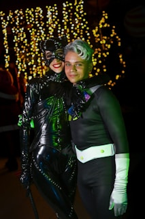 Two people dressed in costumes pose together. The person on the left wears a black, shiny, full-body catsuit complete with a mask and cat ears. The person on the right is in a black outfit with a white belt and gloves, featuring a distinctive hairstyle with silver and green color. They are standing in front of a background adorned with warm, blurry string lights.