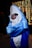 A person wearing a large, plush shark costume poses with their arms crossed. The costume is bright blue with a white underbelly and features large eyes and a wide mouth. The person inside the costume is wearing dark sunglasses, adding a humorous touch. In the background, decorative string lights create a festive atmosphere, with other blurred figures visible.