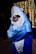 A person wearing a large, plush shark costume poses with their arms crossed. The costume is bright blue with a white underbelly and features large eyes and a wide mouth. The person inside the costume is wearing dark sunglasses, adding a humorous touch. In the background, decorative string lights create a festive atmosphere, with other blurred figures visible.