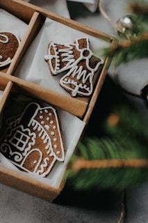A close-up of a box containing intricately decorated gingerbread cookies shaped like a house and a bell with a bow. The cookies are adorned with white icing patterns, giving them a festive appearance. A small portion of a green pine branch is visible in the foreground, adding to the holiday theme.