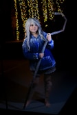 A person with long silver hair dressed in a blue and white patterned robe stands in a dimly lit environment. They are holding a large, ornate staff. In the background, there are yellow fairy lights hanging, creating a warm glow.