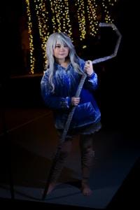 A person with long silver hair dressed in a blue and white patterned robe stands in a dimly lit environment. They are holding a large, ornate staff. In the background, there are yellow fairy lights hanging, creating a warm glow.