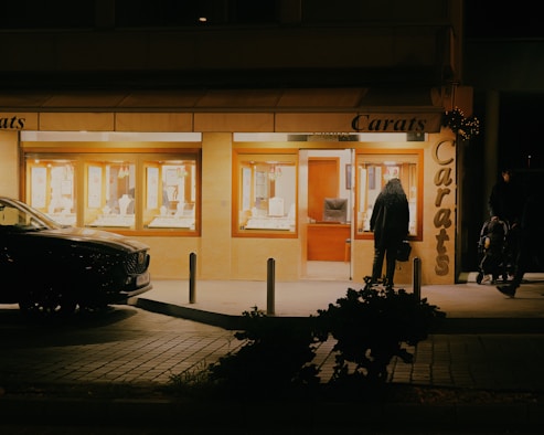 A jewelry store named 'Carats' is illuminated during the night. The storefront features large windows displaying various jewelry pieces under bright lights. A dark-colored car is parked in front of the store. Two people are visible outside the store; one is closer to the entrance while the other is near the right edge of the image, pushing a stroller. The store and surroundings are dimly lit, creating a contrast between the bright store interior and the darker exterior.