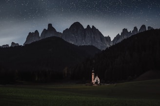 A small church stands isolated in a vast, green meadow surrounded by dark forests. Majestic mountains loom in the background under a sky filled with stars, creating a tranquil and serene atmosphere.