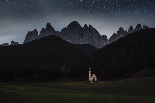 A small church stands isolated in a vast, green meadow surrounded by dark forests. Majestic mountains loom in the background under a sky filled with stars, creating a tranquil and serene atmosphere.