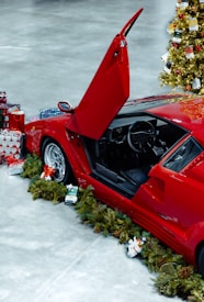 A sporty red car with its door raised is adorned with Christmas decorations. Surrounding the car are various wrapped gifts and green garlands. A Christmas tree with ornaments and lights stands nearby, adding to the festive atmosphere.