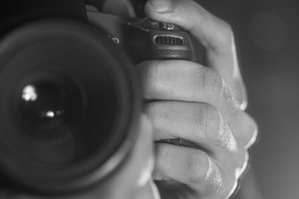 A close-up black and white photograph of a person's hand gripping a camera, focusing on the textured surface of the fingers and the details of the camera's body. The lens is prominent in the foreground, with light reflecting off its surface.