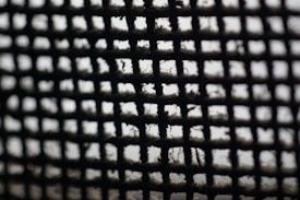 A close-up view of a wire mesh screen with a grid pattern. The mesh appears to have collected dust or lint, and the focus is soft, creating a moody and abstract effect.