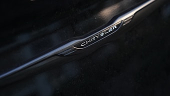 A close-up of a metallic logo featuring the brand name 'Chrysler' on a dark surface. The emblem is sleek with a polished, reflective finish and embossed lettering.