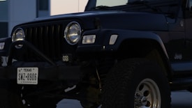 A black Jeep with a Texas license plate is parked, showing a close-up view of the front part. The vehicle has prominent round headlights, a distinctive grille, and rugged tires. The background includes part of a building.