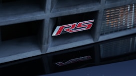 A close-up view of a car's grille featuring a red RS badge set against a textured black background with a reflection at the bottom. The grille is framed by a headlight with a textured glass cover on the right side.