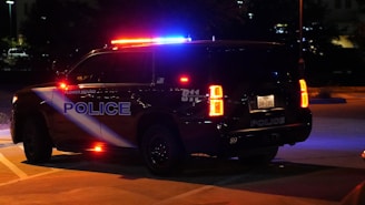 A Flower Mound police SUV is parked with its red and blue emergency lights on in a dimly lit parking lot. The vehicle's markings and license plate are visible, and it is surrounded by darkness with some faintly lit buildings in the background.