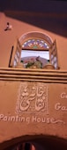 An arched window with open wooden shutters, featuring multi-colored stained glass at the top. Below the window, intricate decorative plasterwork displays script, and the words 'Painting House' are inscribed on the wall. A potted plant with pink and red flowers is visible on the window ledge.