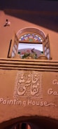 An arched window with open wooden shutters, featuring multi-colored stained glass at the top. Below the window, intricate decorative plasterwork displays script, and the words 'Painting House' are inscribed on the wall. A potted plant with pink and red flowers is visible on the window ledge.