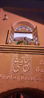 An arched window with open wooden shutters, featuring multi-colored stained glass at the top. Below the window, intricate decorative plasterwork displays script, and the words 'Painting House' are inscribed on the wall. A potted plant with pink and red flowers is visible on the window ledge.