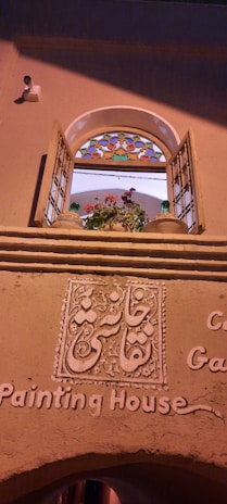 An arched window with open wooden shutters, featuring multi-colored stained glass at the top. Below the window, intricate decorative plasterwork displays script, and the words 'Painting House' are inscribed on the wall. A potted plant with pink and red flowers is visible on the window ledge.