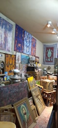 A cluttered room filled with various art pieces, including framed paintings stacked against the walls, a tapestry hanging on a wall, and numerous decorative items like a teapot, baskets, and a vintage radio. The walls are adorned with colorful abstract paintings in blues, reds, and purples, while a large rug with intricate patterns hangs on the wall.