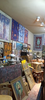A cluttered room filled with various art pieces, including framed paintings stacked against the walls, a tapestry hanging on a wall, and numerous decorative items like a teapot, baskets, and a vintage radio. The walls are adorned with colorful abstract paintings in blues, reds, and purples, while a large rug with intricate patterns hangs on the wall.