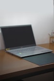A laptop with a black screen sits open on a wooden desk. The laptop is positioned slightly to the left with a black mouse pad partially beneath it. In the background, there are some writing utensils in a holder.
