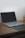 A laptop with a black screen sits open on a wooden desk. The laptop is positioned slightly to the left with a black mouse pad partially beneath it. In the background, there are some writing utensils in a holder.