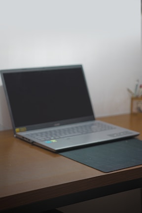 A laptop with a black screen sits open on a wooden desk. The laptop is positioned slightly to the left with a black mouse pad partially beneath it. In the background, there are some writing utensils in a holder.