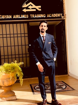 A man in a formal dark suit stands confidently outside a building with a sign that reads 'Libyan Airlines Training Academy'. He is positioned in front of a door, with a decorative plant in a pot nearby.