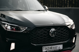 A sleek black car is prominently displayed, featuring a modern and aggressive front design with a distinctive hexagonal grille bearing the MG logo. The headlights are stylish with angular shapes, and the surface reflects light, highlighting the car's glossy finish.