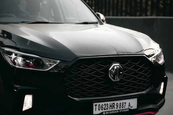 A sleek black car is prominently displayed, featuring a modern and aggressive front design with a distinctive hexagonal grille bearing the MG logo. The headlights are stylish with angular shapes, and the surface reflects light, highlighting the car's glossy finish.