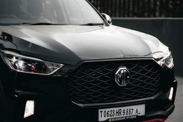 A sleek black car is prominently displayed, featuring a modern and aggressive front design with a distinctive hexagonal grille bearing the MG logo. The headlights are stylish with angular shapes, and the surface reflects light, highlighting the car's glossy finish.