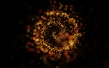 A swirling pattern of glowing, bokeh-like light particles in hues of gold and red against a dark background creates a mesmerizing and dynamic visual effect.