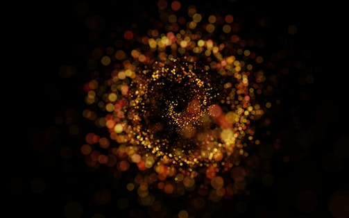 A swirling pattern of glowing, bokeh-like light particles in hues of gold and red against a dark background creates a mesmerizing and dynamic visual effect.