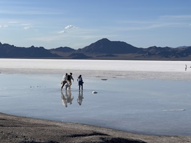 A vast salt flat stretches into the distance, reflecting the clear blue sky and surrounding mountains. A group of people walks across the shallow layer of water covering the salt surface, creating reflections of their figures. Mountains form a dark silhouette against the serene backdrop.