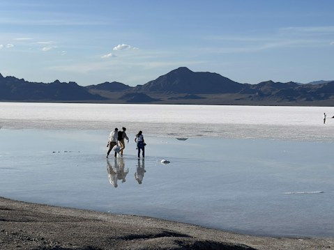 A vast salt flat stretches into the distance, reflecting the clear blue sky and surrounding mountains. A group of people walks across the shallow layer of water covering the salt surface, creating reflections of their figures. Mountains form a dark silhouette against the serene backdrop.