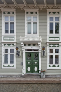 A charming facade of a historic building features symmetrical green and white wooden panels with ornate decorative elements. The central focus is a pair of green doors adorned with intricate carvings and glass panes, flanked by lantern-style wall lamps. Above, white decorative trims and molding add elegance to the windows. At the base of the steps, a white pot filled with red flowers adds a touch of color.