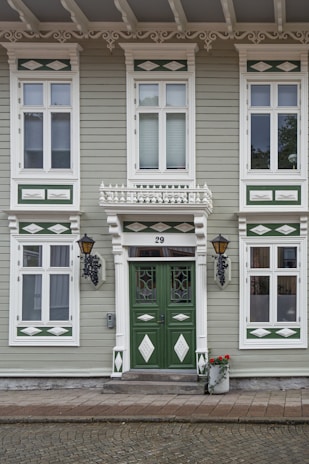 A charming facade of a historic building features symmetrical green and white wooden panels with ornate decorative elements. The central focus is a pair of green doors adorned with intricate carvings and glass panes, flanked by lantern-style wall lamps. Above, white decorative trims and molding add elegance to the windows. At the base of the steps, a white pot filled with red flowers adds a touch of color.